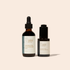Two amber glass skincare bottles from Faithful Skincare on a warm beige background. The taller bottle on the left is Ozonated Cleansing Oil with a black dropper cap, and the shorter bottle on the right is Ozonated Facial Serum with a black pump cap.