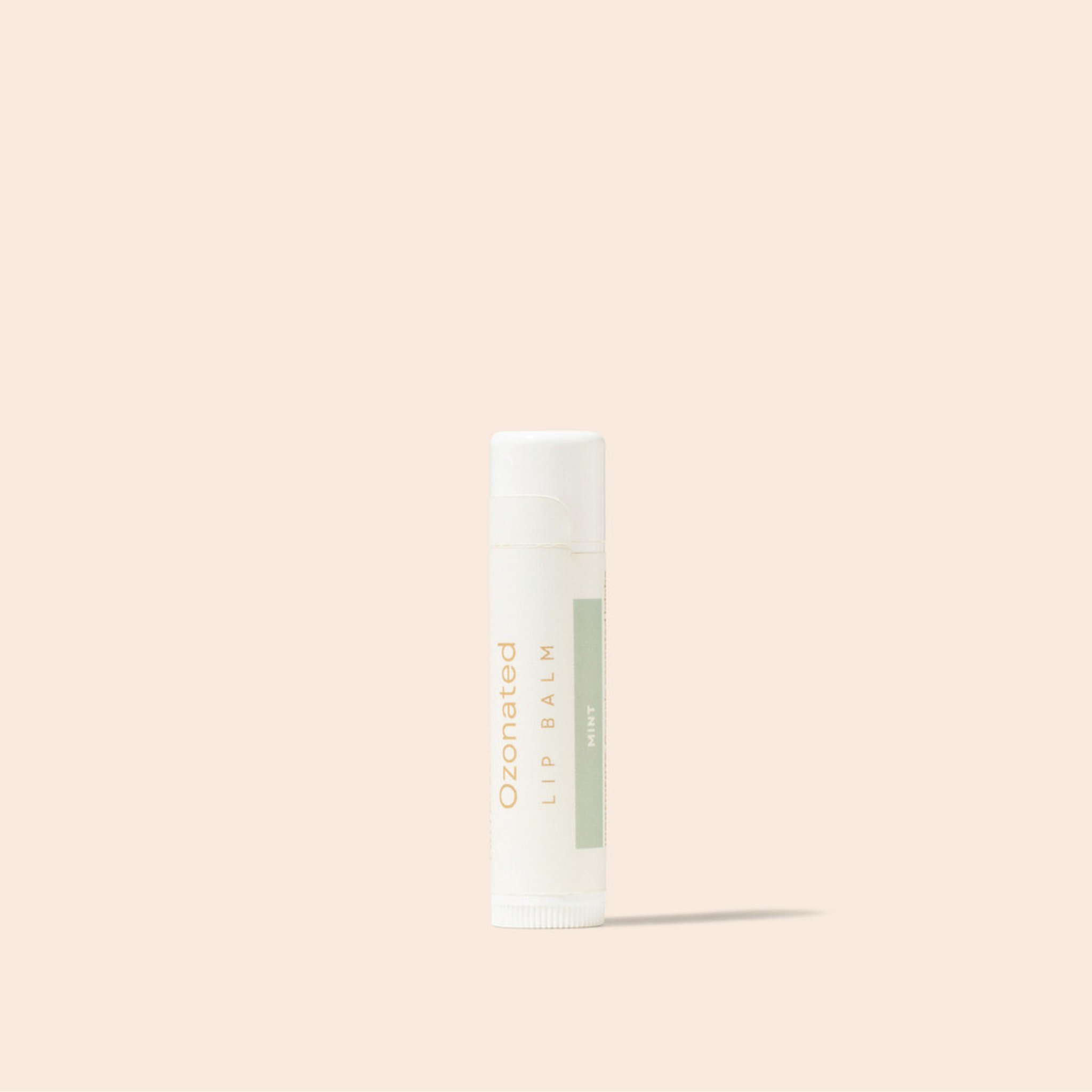 A single tube of ozonated lip balm with a white cap and a white body featuring a light sage green label, standing upright against a solid pale peach-beige background.