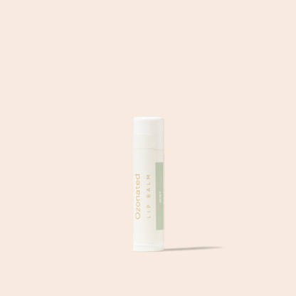 A single tube of ozonated lip balm with a white cap and a white body featuring a light sage green label, standing upright against a solid pale peach-beige background.